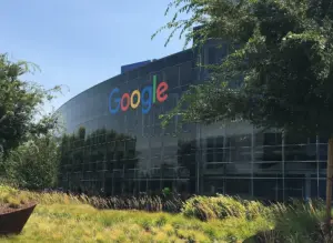 Логото Google върху стъклената фасада на сграда в Googleplex сред дървета.