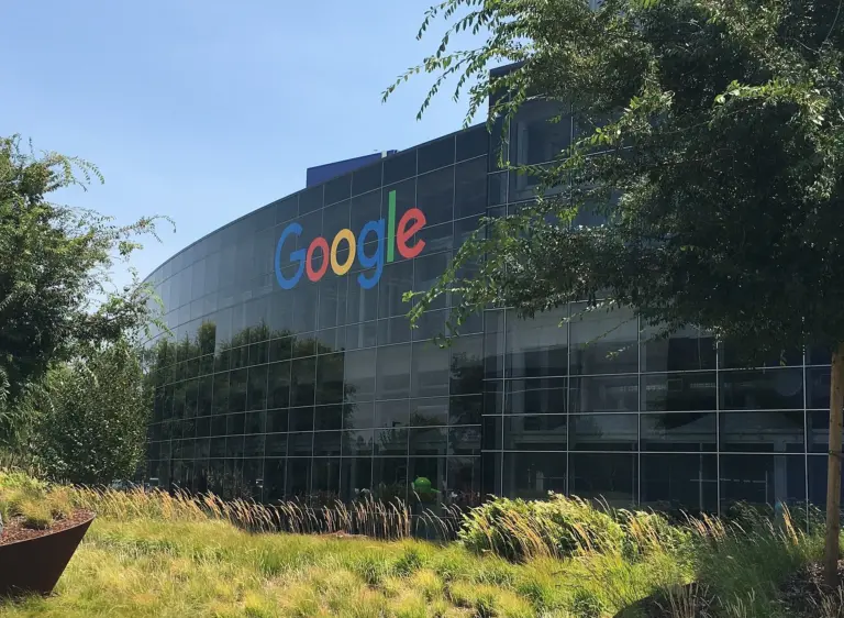 Логото Google върху стъклената фасада на сграда в Googleplex сред дървета.